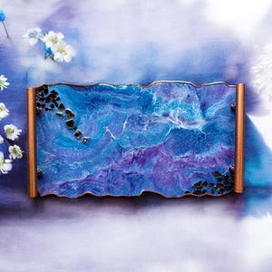 May include: A rectangular resin tray with a wavy edge, featuring a blend of blue and purple hues with a marbled effect. The tray has copper-colored sides and is adorned with black stones. White flowers and blue accents surround the tray.