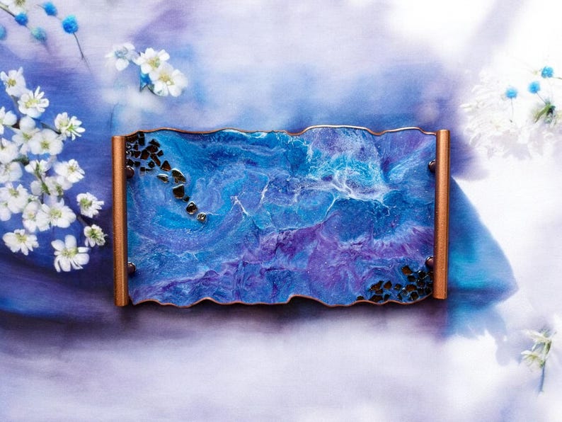May include: A rectangular resin tray with a wavy edge, featuring a blend of blue and purple hues with a marbled effect. The tray has copper-colored sides and is adorned with black stones. White flowers and blue accents surround the tray.