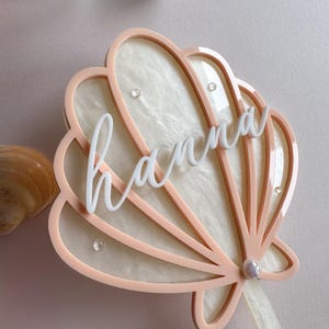 May include: A light pink seashell-shaped cake topper with the name "hanna" in white script. The topper has a pearl accent and a ribbon. Decorative seashells are in the background.