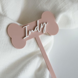 May include: A pink bone-shaped cake topper with the name "Indy" written in white lettering.