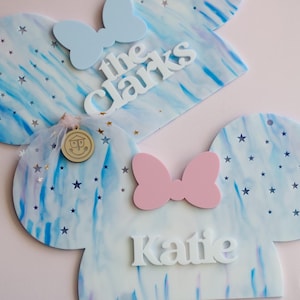 May include: Two blue and white Mickey Mouse ears with stars and a swirl pattern. The ears are personalised with the names "The Clarks" and "Katie".
