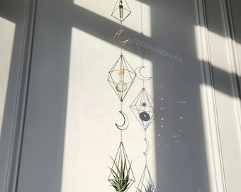 May include: A gold geometric hanging mobile with crystals, air plants, and crescent moons. The mobile is hanging in front of a white wall with shadows from a window.