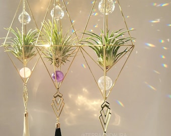 May include: Three hanging geometric air plant holders with gold metal frames, each holding a green air plant. The holders feature crystal accents, decorative tassels, and a chain for hanging. The design is modern and decorative.
