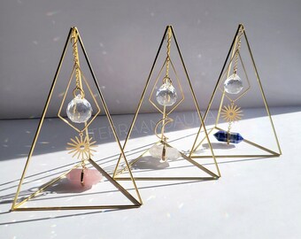 May include: Three gold triangular stands with crystal prisms hanging from the top. Each stand has a different crystal at the bottom: a pink crystal, a white crystal, and a blue crystal.