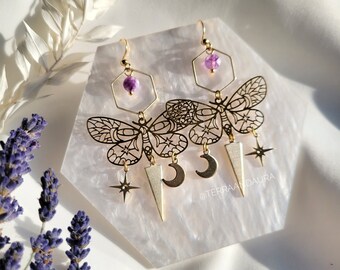 May include: Gold butterfly earrings with amethyst gemstone accents, crescent moon and star charms, and a geometric design. The earrings are displayed on a white hexagonal surface. @terraandaura