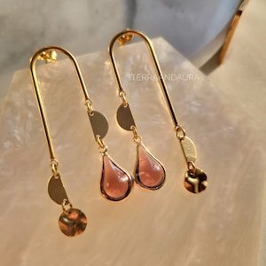 Peut inclure: Une paire de boucles d'oreilles en or avec une pierre rose en forme de larme et des formes géométriques. Les boucles d'oreilles sont suspendues à une barre en or.