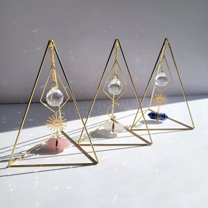 May include: Three gold triangular stands with crystal prisms hanging from the top. Each stand has a different crystal at the bottom: a pink crystal, a white crystal, and a blue crystal.