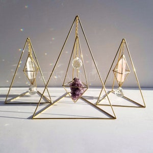 May include: Three gold geometric crystal holders with hanging crystals. The holders are triangular in shape and have a diamond-shaped opening in the centre. The crystals are a clear quartz point, a pink quartz point, and a purple amethyst cluster.