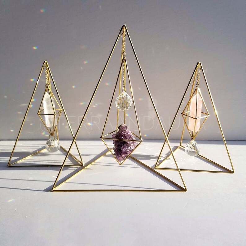 May include: Three gold geometric crystal holders with hanging crystals. The holders are triangular in shape and have a diamond-shaped opening in the centre. The crystals are a clear quartz point, a pink quartz point, and a purple amethyst cluster.