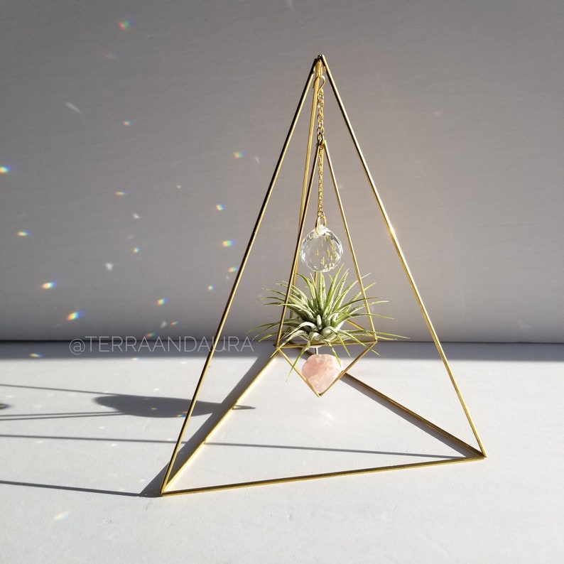 May include: A gold geometric pyramid stand with a hanging crystal prism, a pink heart-shaped crystal, and a small air plant. The stand is on a white surface with a shadow cast by the stand.
