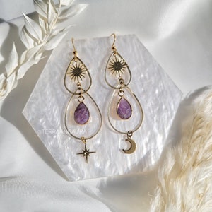 Pode incluir: Brincos de ouro com uma pedra roxa, um sol, uma lua e um amuleto de estrela. Os brincos estão sobre um fundo branco.