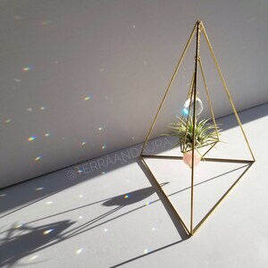 May include: A gold metal geometric pyramid stand with a crystal prism hanging from the top. A small air plant is potted in a pink planter and sits inside the pyramid. The stand casts rainbow reflections on the white surface.