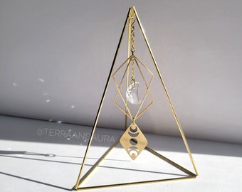 May include: A gold metal pyramid stand with a crystal moon and moon phases charm hanging from it. The charm is attached to the stand with a gold chain.