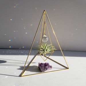 May include: A gold geometric pyramid stand with a hanging crystal prism and a small air plant. A purple amethyst cluster sits on the base of the stand.
