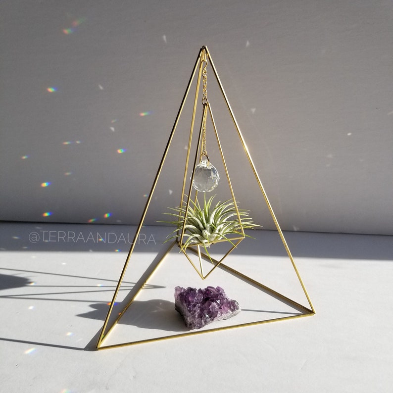 May include: A gold geometric pyramid stand with a hanging crystal prism and a small air plant. A purple amethyst cluster sits on the base of the stand.