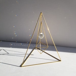 May include: A gold metal pyramid with a crystal hanging from the top. The crystal is suspended by a chain and is reflecting light.