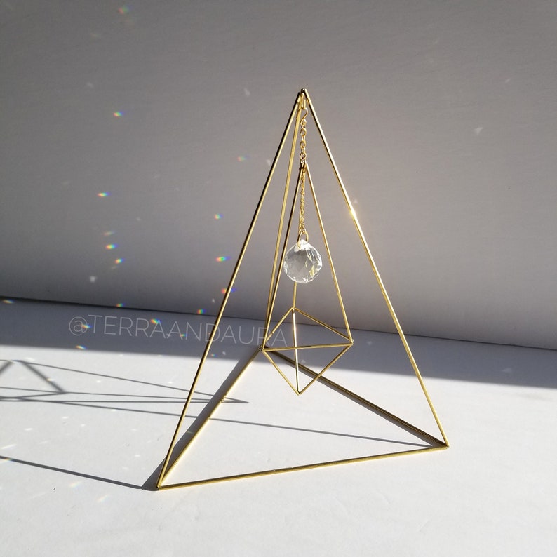 May include: A gold metal pyramid with a crystal hanging from the top. The crystal is suspended by a chain and is reflecting light.