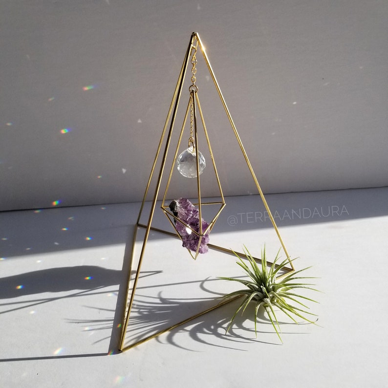 May include: A gold metal pyramid stand with a crystal ball hanging from the top and a purple amethyst crystal inside. The stand is on a white surface with a green air plant in the foreground. The text "@TERRAANDLAURA" is visible in the background.