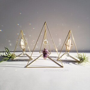 May include: Three gold geometric crystal display stands featuring amethyst, clear quartz, and a clear crystal ball. The stands are pyramid-shaped with a diamond-shaped opening in the centre. The stands are on a white surface with the text "@terraaandaaura" at the bottom.