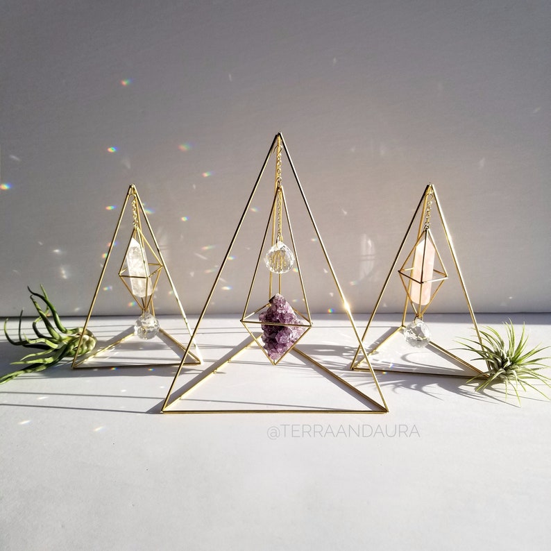 May include: Three gold geometric crystal display stands featuring amethyst, clear quartz, and a clear crystal ball. The stands are pyramid-shaped with a diamond-shaped opening in the centre. The stands are on a white surface with the text "@terraaandaaura" at the bottom.