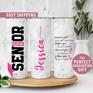 May include: Two white marble-patterned stainless steel tumblers with pink and black text. The first tumbler says "Senior 2023" with a graduation cap. The second tumbler says "Jessica" and has a quote about memories, dreams, and love. The tumblers are on a wooden surface.
