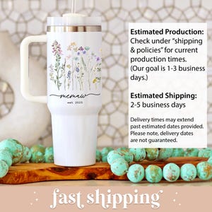 May include: White tumbler with a handle, featuring a floral design in pastel colors. The tumbler has the text "memaw est 2025". The tumbler is on a wooden surface with turquoise beads.