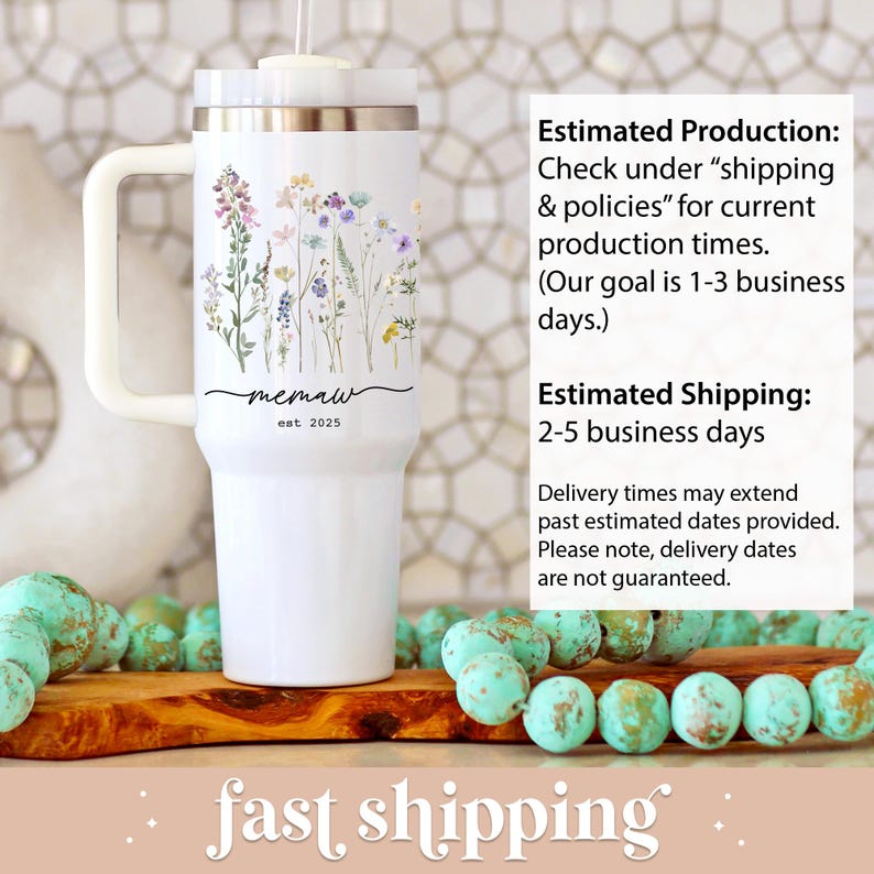 May include: White tumbler with a handle, featuring a floral design in pastel colors. The tumbler has the text "memaw est 2025". The tumbler is on a wooden surface with turquoise beads.