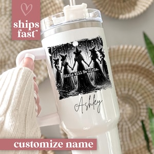 May include: A white tumbler with a handle and a silver lid. The tumbler features a black and white illustration of witches holding hands with the text "Girls Will Be Girls" and a custom name. The tumbler has a straw.