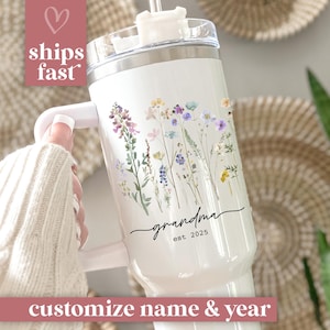 May include: White insulated tumbler with a handle, featuring a floral design in purple, yellow, and blue. The word "grandma" is written in script, with "est. 2025" below. The text "customize name & year" is at the bottom.