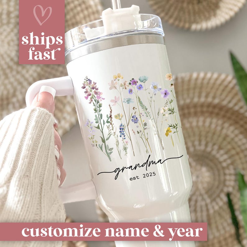 May include: White insulated tumbler with a handle, featuring a floral design in purple, yellow, and blue. The word "grandma" is written in script, with "est. 2025" below. The text "customize name & year" is at the bottom.