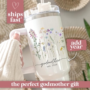 May include: White tumbler with a floral design and the word "Godmother" in black script. The tumbler also has the text "est. 2026". The tumbler has a white handle and a clear lid with a straw.