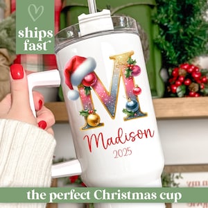 May include: White insulated tumbler with a handle, featuring a large letter "M" decorated with Christmas ornaments and a Santa hat. The name "Madison" and the year "2025" are printed below the letter. The text "ships fast" and "the perfect Christmas cup" are also visible.