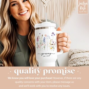 May include: White travel mug with a floral design and the word "granny" in script, with "est. 2025" below. The mug has a handle and a white lid with a straw. The mug is held by a person.