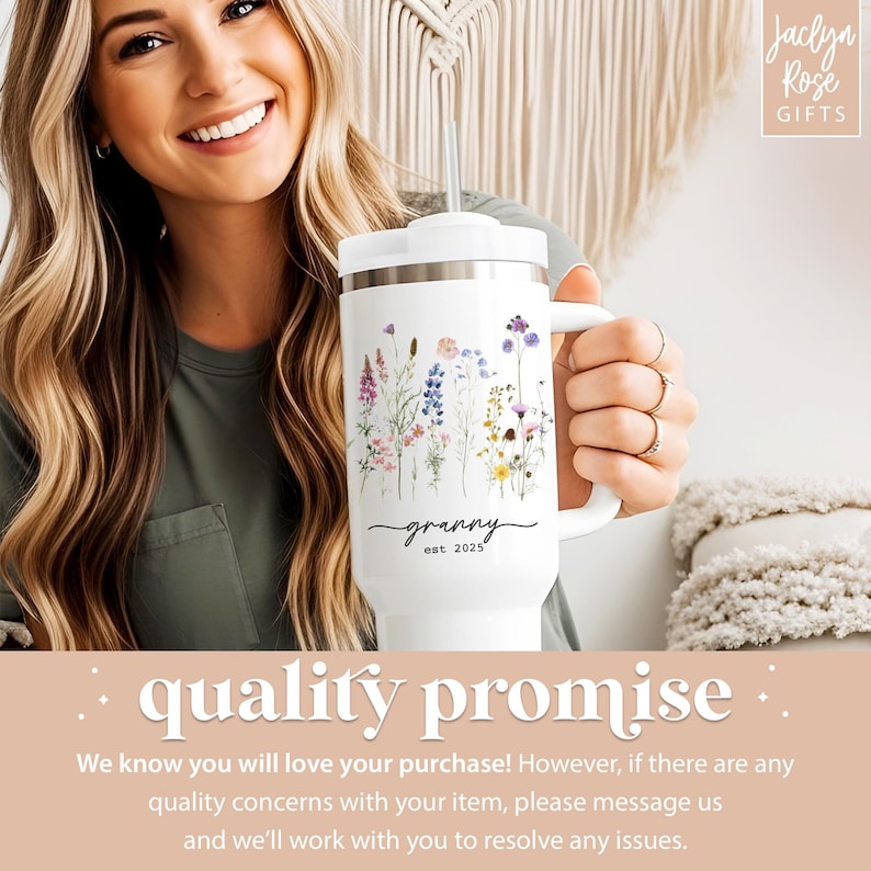 May include: White travel mug with a floral design and the word "granny" in script, with "est. 2025" below. The mug has a handle and a white lid with a straw. The mug is held by a person.