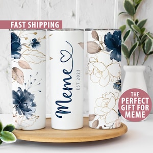 May include: Three white tumblers with a floral design in shades of blue and gold. The middle tumbler has the word "meme" written in blue with a heart and the text "EST 2023".