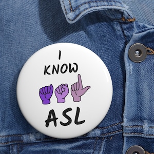 Könnte beinhalten: Weißer Knopf mit dem Text "I KNOW ASL" in Schwarz. Die Buchstaben "ASL" werden durch drei violette und rosa Handzeichen dargestellt.