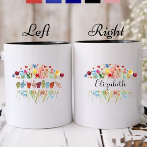 May include: Two white ceramic mugs with black rims and handles. The mug on the left has a floral design with a sign language interpretation of the name "Elizabeth". The mug on the right has a floral design with the name "Elizabeth" written in cursive. The mugs are available in multiple handle colors.
