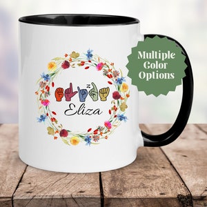 May include: White ceramic mug with a black handle and rim. The mug features a floral wreath design with colorful flowers and the name "Eliza" in script. The mug also displays the name "Eliza" in American Sign Language.