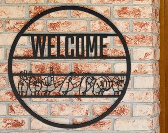 May include: A black metal welcome sign with a circular frame is mounted on a brick wall. The word "WELCOME" is displayed across the top, with a decorative design below. A black outdoor light and a wooden door are also visible.
