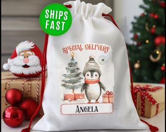 May include: White drawstring Christmas gift bag with a penguin illustration wearing a hat and scarf. The bag has the text "Special Delivery" and the name "Angela". A green circle with "Ships Fast" is visible.