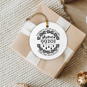 May include: A white, round holiday ornament with the text "There's no place like home 99201 for the holidays" in black script. The ornament has a gold string for hanging and is placed on a wrapped gift.