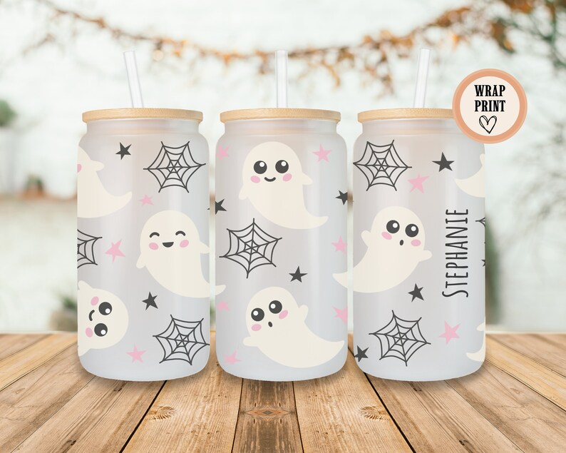 May include: Three glass beverage cups with a Halloween theme. The cups feature a wrap-around print of smiling ghosts, spiderwebs, and stars in black and pink on a frosted background. Each cup has a bamboo lid and a straw.