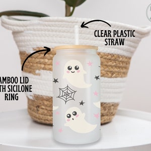 May include: A frosted glass tumbler with a bamboo lid and clear plastic straw. The tumbler features a repeating pattern of cute ghost illustrations, pink stars, black stars, and spiderwebs. The tumbler is designed for cold beverages.