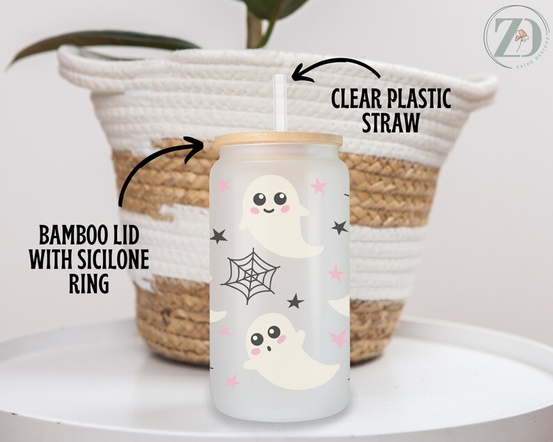 May include: A frosted glass tumbler with a bamboo lid and clear plastic straw. The tumbler features a repeating pattern of cute ghost illustrations, pink stars, black stars, and spiderwebs. The tumbler is designed for cold beverages.