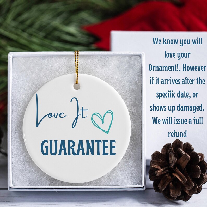May include: A white, round ceramic ornament with the words "Love It" and "GUARANTEE" in blue script. A teal heart graphic is next to "It". The ornament is in a white box with a gold string for hanging.