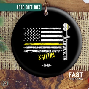 May include: Black round ornament featuring a distressed American flag design with a yellow stripe. The ornament includes the name "KAITUN" in yellow, the year "-2025-", and the text "911 DISPATCHER". A heart and heartbeat graphic is also present.