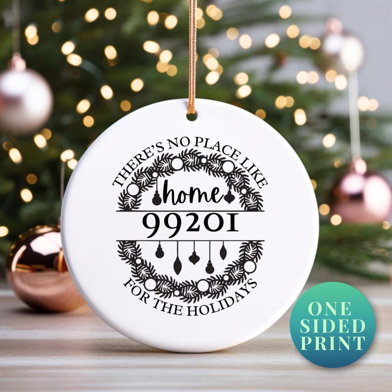 May include: A white ceramic Christmas ornament with the text "THERE'S NO PLACE LIKE home FOR THE HOLIDAYS" and the zip code 99201. The ornament has a gold string for hanging and a green and blue circle that says "ONE SIDED PRINT".