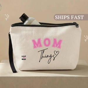 May include: A cream-colored canvas cosmetic bag with a black zipper and handle. The bag features the words "MOM Things" in pink and black script, with the names "Lilly Noah" below. A bottle is visible inside.