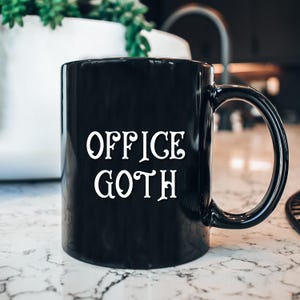May include: Black ceramic mug with the words "OFFICE GOTH" in white, stylized lettering. The mug has a curved handle and sits on a marble-patterned surface. A plant in a white pot is visible in the background.