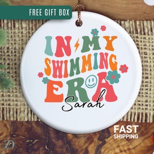 May include: A white ceramic ornament with the text "IN MY SWIMMING ERA" in retro colors, with the name "Sarah" below. The ornament has a gold string for hanging. The words "FREE GIFT BOX" and "FAST SHIPPING" are also visible.